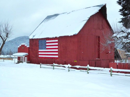 A red barn covered in snow features a large American flag painted on its side, set against a winter landscape.