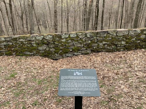A sign on a metal stand in front of a moss-covered stone wall, surrounded by a forest of bare trees and fallen leaves.