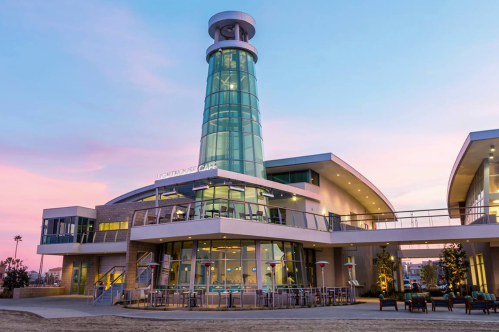 Modern building with a glass tower, outdoor seating, and a colorful sunset sky in the background.