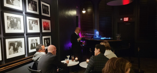A dimly lit bar with framed photos on the wall, a pianist performing, and patrons enjoying drinks at tables.