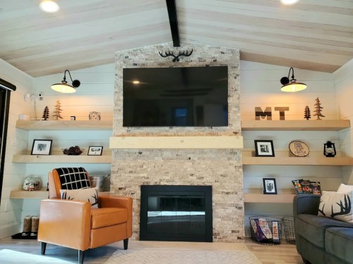 Cozy living room with a stone fireplace, mounted TV, wooden shelves, and stylish furniture. Warm, inviting decor.