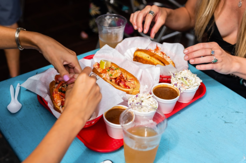 Two people enjoy lobster rolls, coleslaw, and drinks on a vibrant blue table.