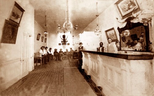 A vintage bar scene with patrons seated at tables and a bartender behind the counter, featuring period decor and lighting.