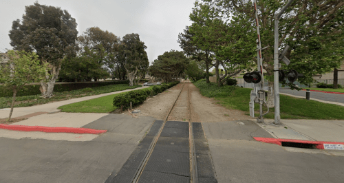A view of a railway crossing with tracks, surrounded by trees and greenery, on a cloudy day.