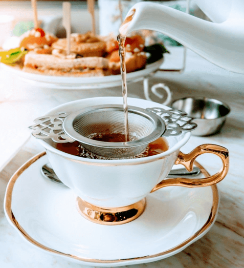 A teapot pours tea into a delicate white cup with a gold rim, featuring a metal strainer, with snacks in the background.