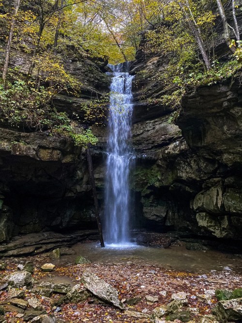 A serene waterfall cascades down rocky cliffs, surrounded by autumn foliage and a tranquil pool below.