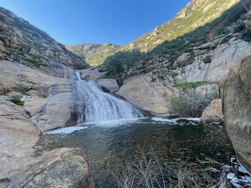 A serene waterfall cascades over rocky terrain into a tranquil pool, surrounded by lush greenery and mountains.