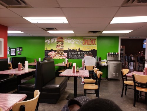 Interior of a casual restaurant with green walls, checkered tables, and a large mural promoting a burger challenge.