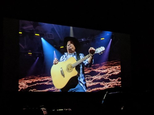 A musician in a cowboy hat plays an acoustic guitar on stage, with colorful lights and a dramatic backdrop.