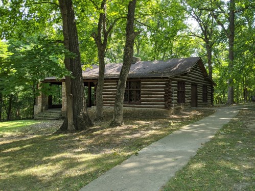 A rustic log cabin surrounded by trees, with a stone pathway leading to the entrance.