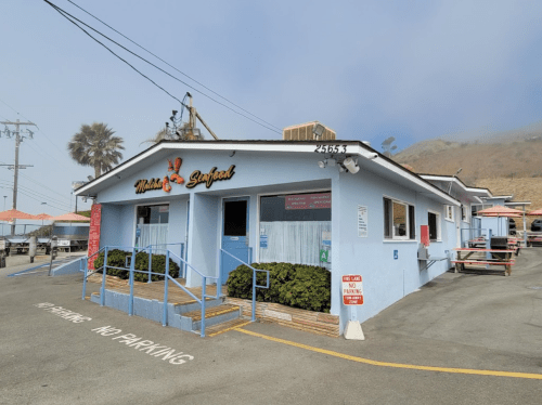 A blue seafood restaurant with a sign, outdoor seating, and no parking signs in a coastal area.
