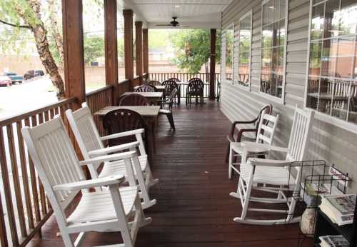 A porch with wooden flooring, featuring rocking chairs and tables, surrounded by greenery and a view of the street.