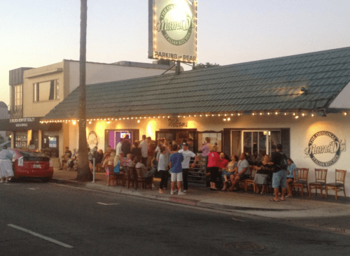 A bustling restaurant exterior with a crowd gathered outside, string lights illuminating the entrance.