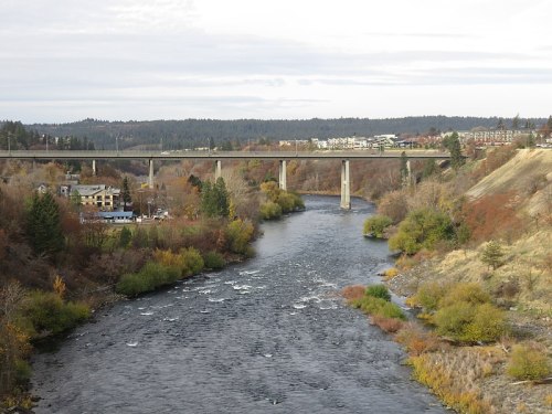 A river flows beneath a bridge, surrounded by trees and hills, with buildings visible in the background.