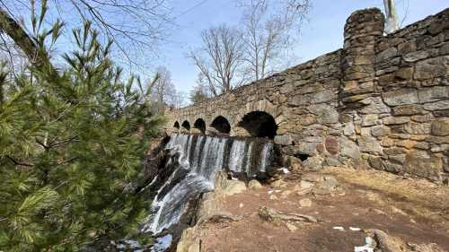 A stone bridge with arches over a waterfall, surrounded by trees and rocky terrain on a clear day.