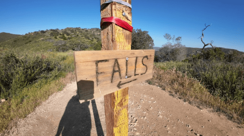 Wooden signpost pointing left towards "Falls," surrounded by greenery and a clear blue sky.