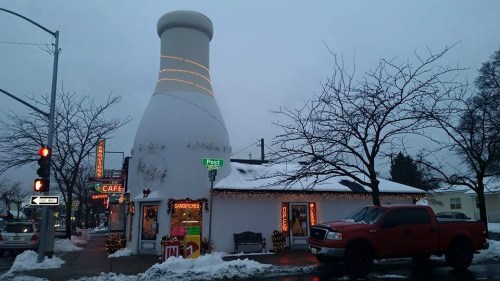 A whimsical building shaped like a milk bottle, decorated for winter, with a red truck parked outside. Snow covers the ground.