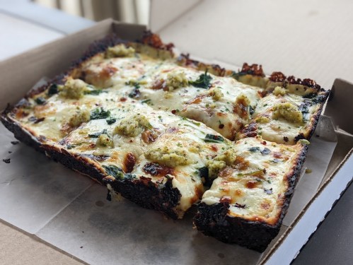 A rectangular pizza with a crispy crust, topped with melted cheese, spinach, and dollops of green sauce, in a pizza box.