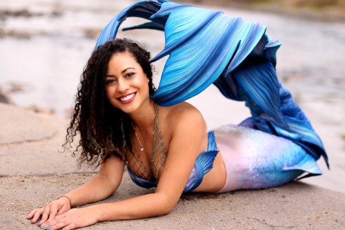 A smiling woman in a colorful mermaid costume poses on a rocky shore, with flowing blue and pink tail details.