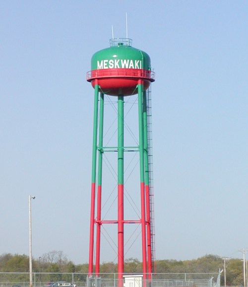 A tall water tower painted in red and green, labeled "MESKWAKI," against a clear blue sky.