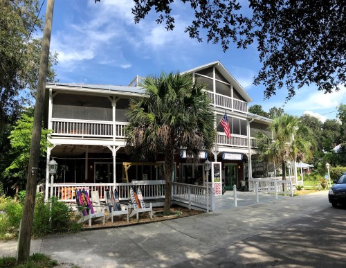 A two-story building with a porch, palm trees, and an American flag, set in a sunny, green environment.