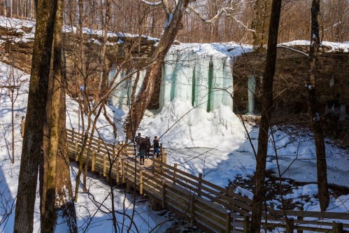 A snowy landscape with a frozen waterfall and people standing on a wooden bridge surrounded by trees.