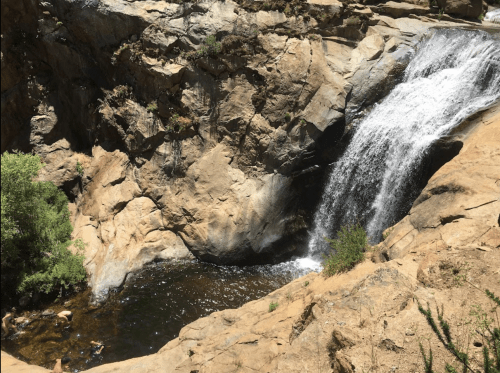 A waterfall cascades over rocky cliffs into a natural pool, surrounded by greenery and people enjoying the water.