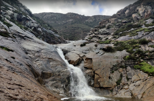 A waterfall cascading down rocky terrain, surrounded by lush greenery and mountains under a cloudy sky.