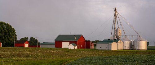 A rural farm scene featuring red barns, silos, and green fields under a cloudy sky.