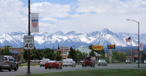 A scenic view of a highway with cars, shops, and snow-capped mountains in the background under a cloudy sky.