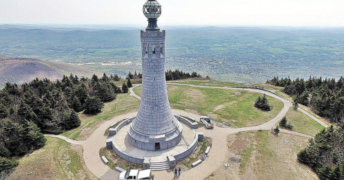 A tall stone tower with a glass dome, surrounded by green hills and trees, under a clear sky.