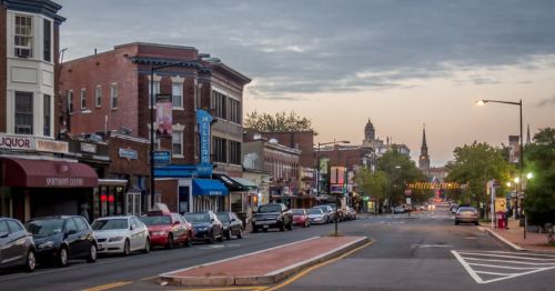 A quiet street lined with shops and parked cars, under a cloudy sky at dawn.