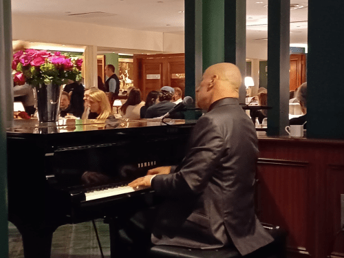 A musician plays piano in a lively restaurant, with guests enjoying their meals in the background.