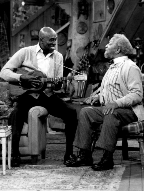 Two elderly men sit in a living room; one plays a ukulele while the other listens, both smiling and engaged.