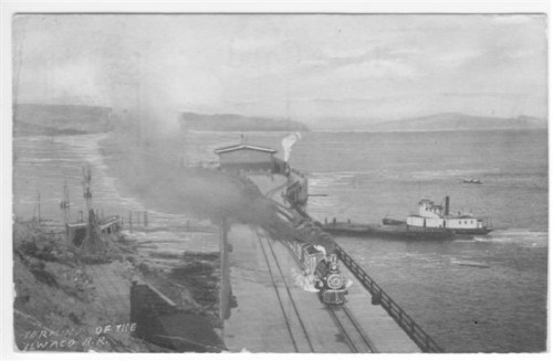 Black and white image of a train arriving at a dock, with a steamboat nearby and a scenic waterfront in the background.