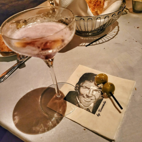 A cocktail in a coupe glass beside a napkin featuring a black-and-white photo of a man and two olives on a skewer.