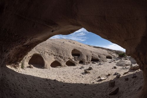 View from inside a cave, showing sandy ground and rock formations under a blue sky with scattered clouds.