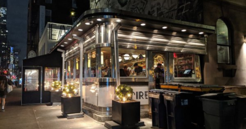 A brightly lit diner at night, featuring a retro design and outdoor seating, surrounded by city lights.
