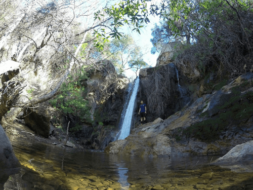 A person stands near a waterfall surrounded by lush greenery and rocky terrain, reflecting in a calm pool of water.