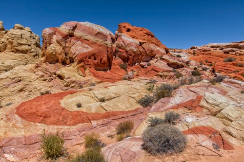 Colorful rock formations with red, orange, and yellow stripes under a clear blue sky, surrounded by sparse vegetation.