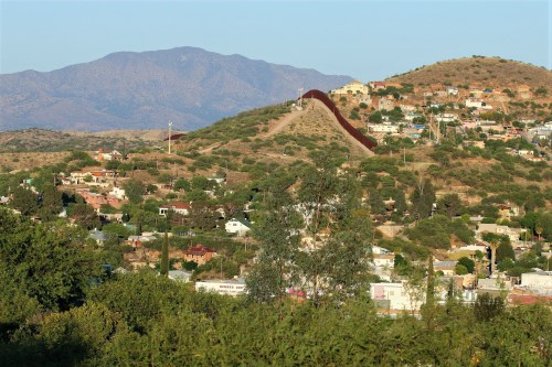 A view of a mountainous landscape with a border wall separating two communities, surrounded by greenery and homes.