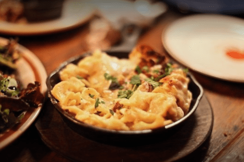 A close-up of a cheesy pasta dish in a small skillet, garnished with herbs, on a wooden table.