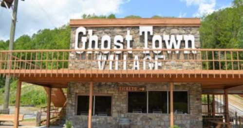 A stone building with "Ghost Town Village" sign and ticket window, surrounded by greenery and blue sky.