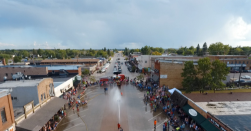 Aerial view of a town parade with a crowd gathered, fire trucks spraying water down the street under a cloudy sky.
