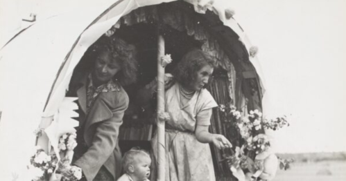 Two women and a baby peek out from a decorated wagon, surrounded by flowers and festive decorations.