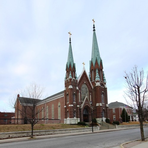 A brick church with tall spires and crosses, set against a cloudy sky, surrounded by bare trees and a quiet street.