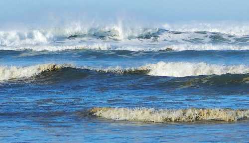 Waves crashing on a sandy beach, with frothy white tops against a deep blue ocean under a clear sky.