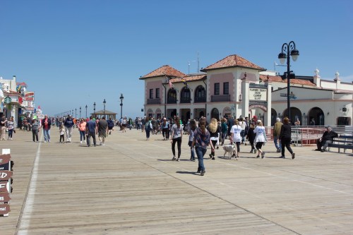 A busy boardwalk with people walking, shops in the background, and a clear blue sky overhead.
