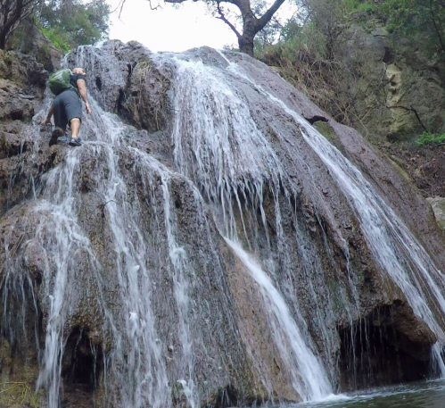 A person climbs a rocky waterfall surrounded by lush greenery. Water cascades down the rocks.