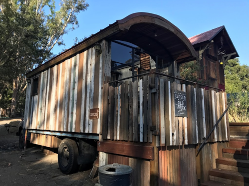 A rustic wooden truck with a curved roof, featuring a "Closed" sign, surrounded by trees and a dirt path.